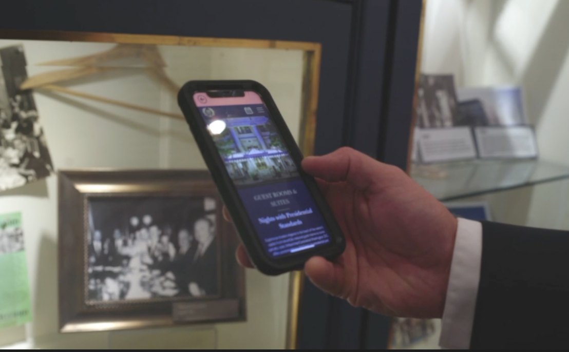 A guest points a phone at a historic display case and is taken to the hotel's guest rooms and suites page